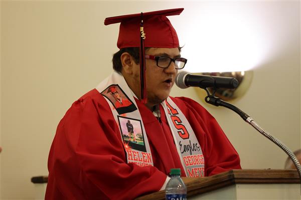 A graduate from the Helen Keller School of Alabama speaks into a microphone from behind a podium. He wears a red cap and gown. He wears a stole decorated with pictures from his time at Helen Keller School of Alabama and text that reads \